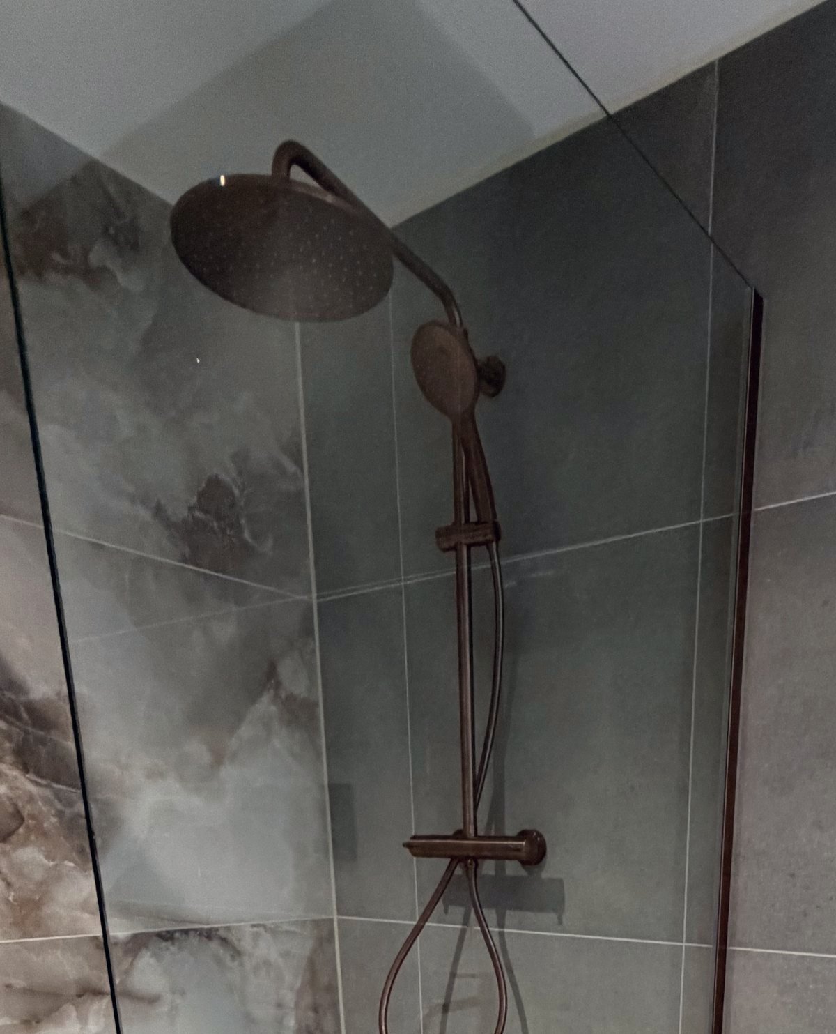 Copper rain shower and handset against grey and marble-effect wall tiles, behind a glass screen